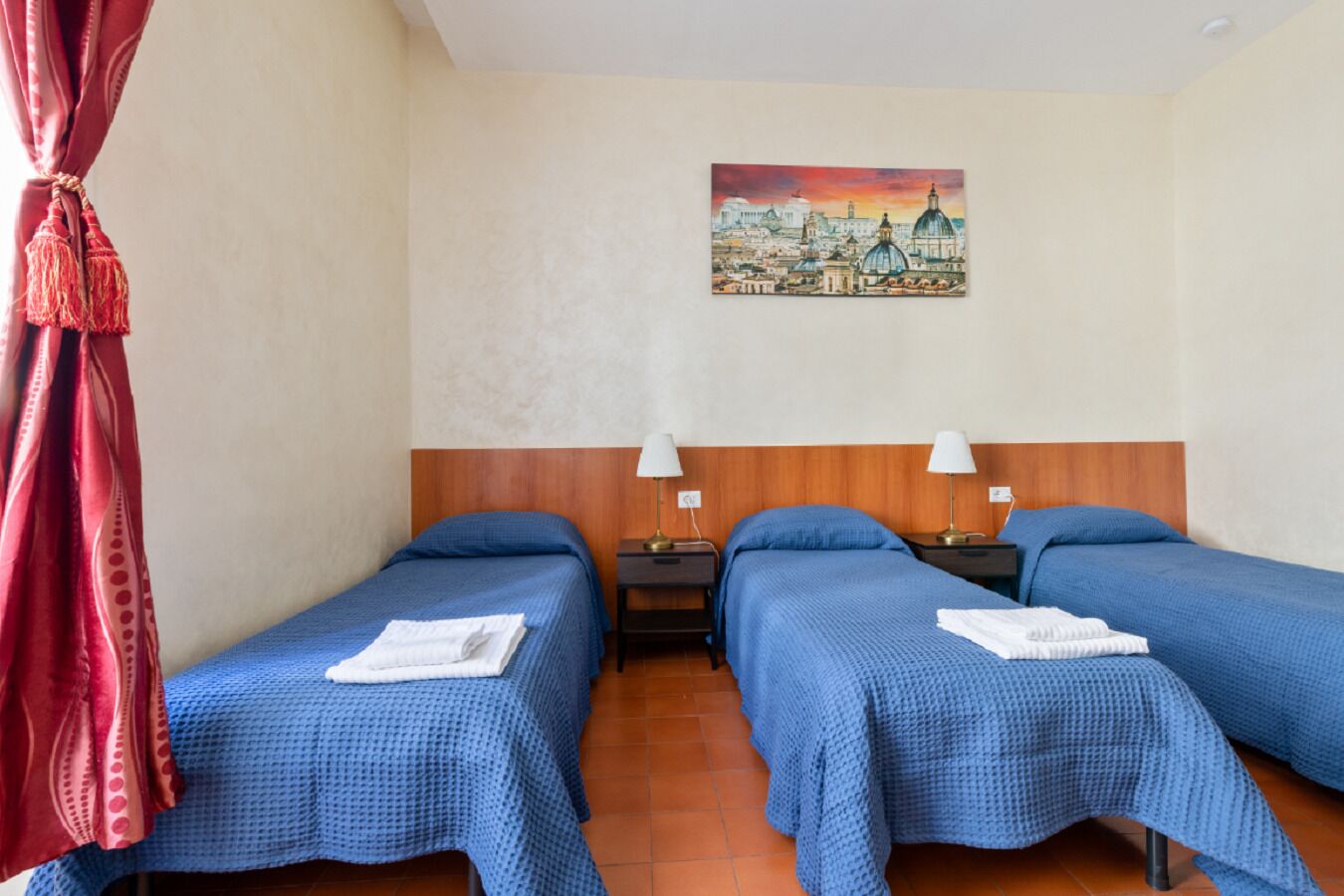 A hotel room with two beds, each with a blue bedspread and white towels, a wooden headboard, a painting of a cityscape on the wall, and a red curtain on the left.