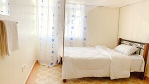 A single bed with a wooden headboard in a room with patterned floor tiles and sheer curtains with blue floral patterns. - NAIROBI SIMBA YOUTH HOSTELS (Syokimau)