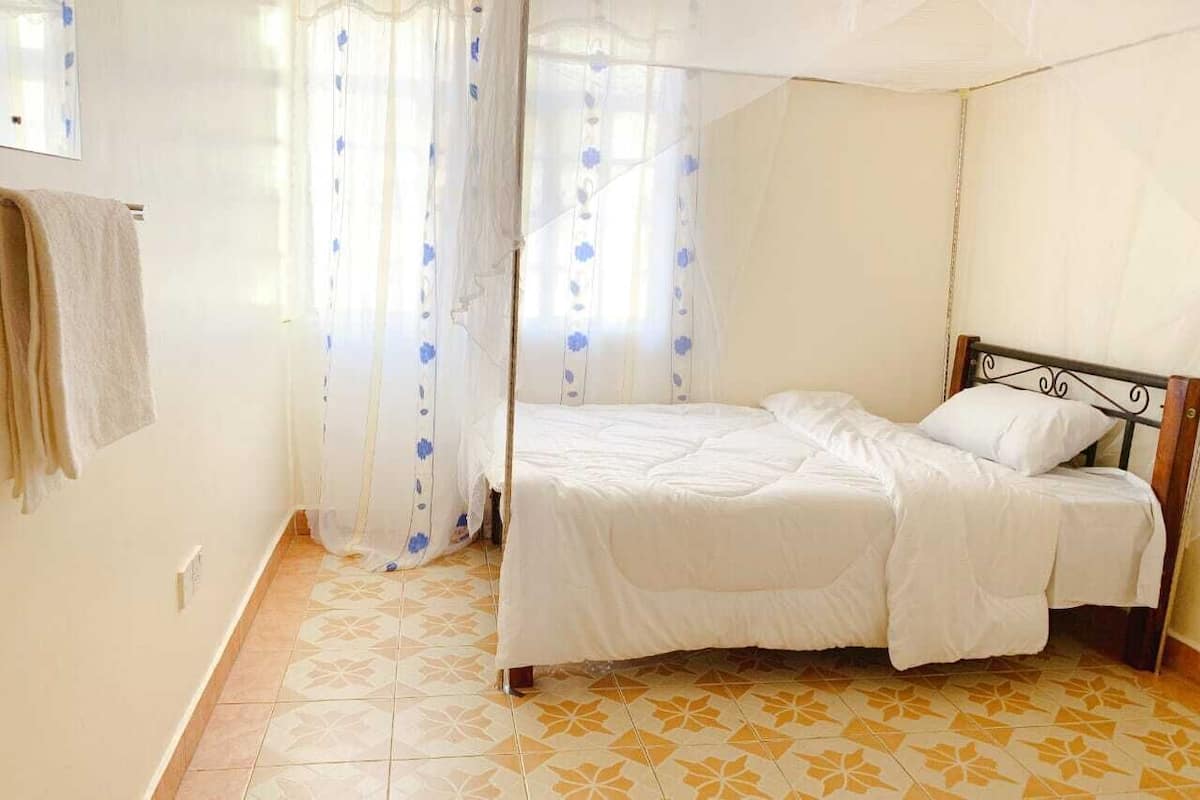 A single bed with a wooden headboard in a room with patterned floor tiles and sheer curtains with blue floral patterns.