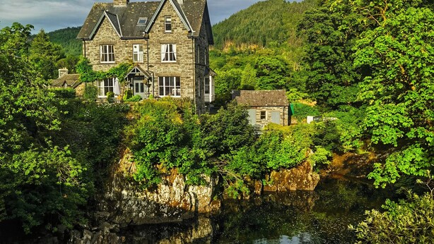 A stone house with a large window and a chimney, surrounded by lush greenery and a small stream. - Tyn Y Fron Country House (Betws-Y-Coed)