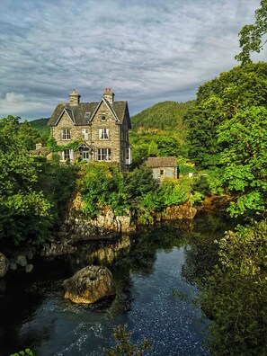 A stone house with a large window and a chimney, surrounded by lush greenery and a small stream. - Tyn Y Fron Country House (Betws-Y-Coed)