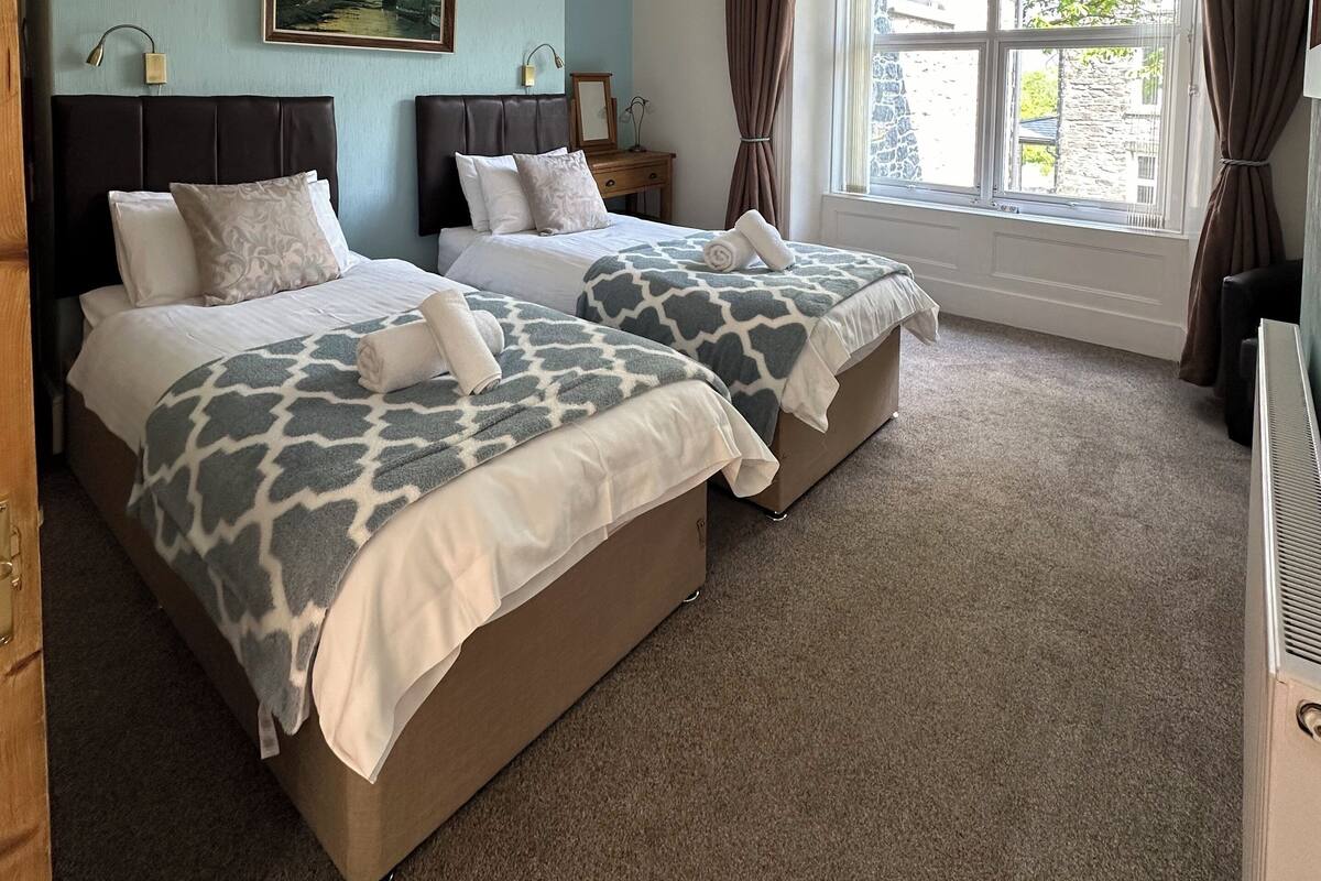 A hotel room with two beds, a ceiling fan, a painting on the wall, and a window with curtains.