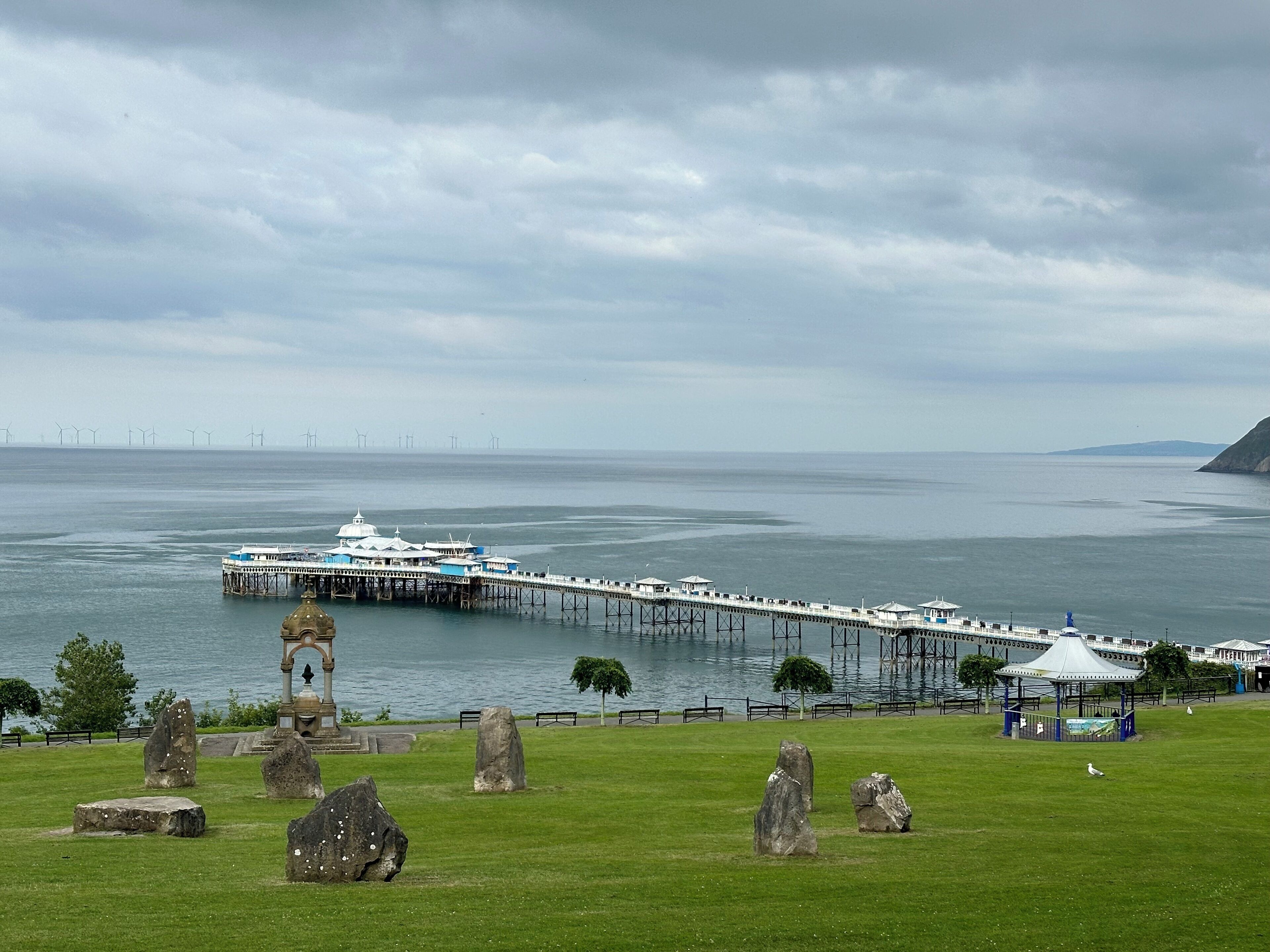 A coastal pier with a gazebo and a lighthouse, surrounded by a grassy area with standing stones.