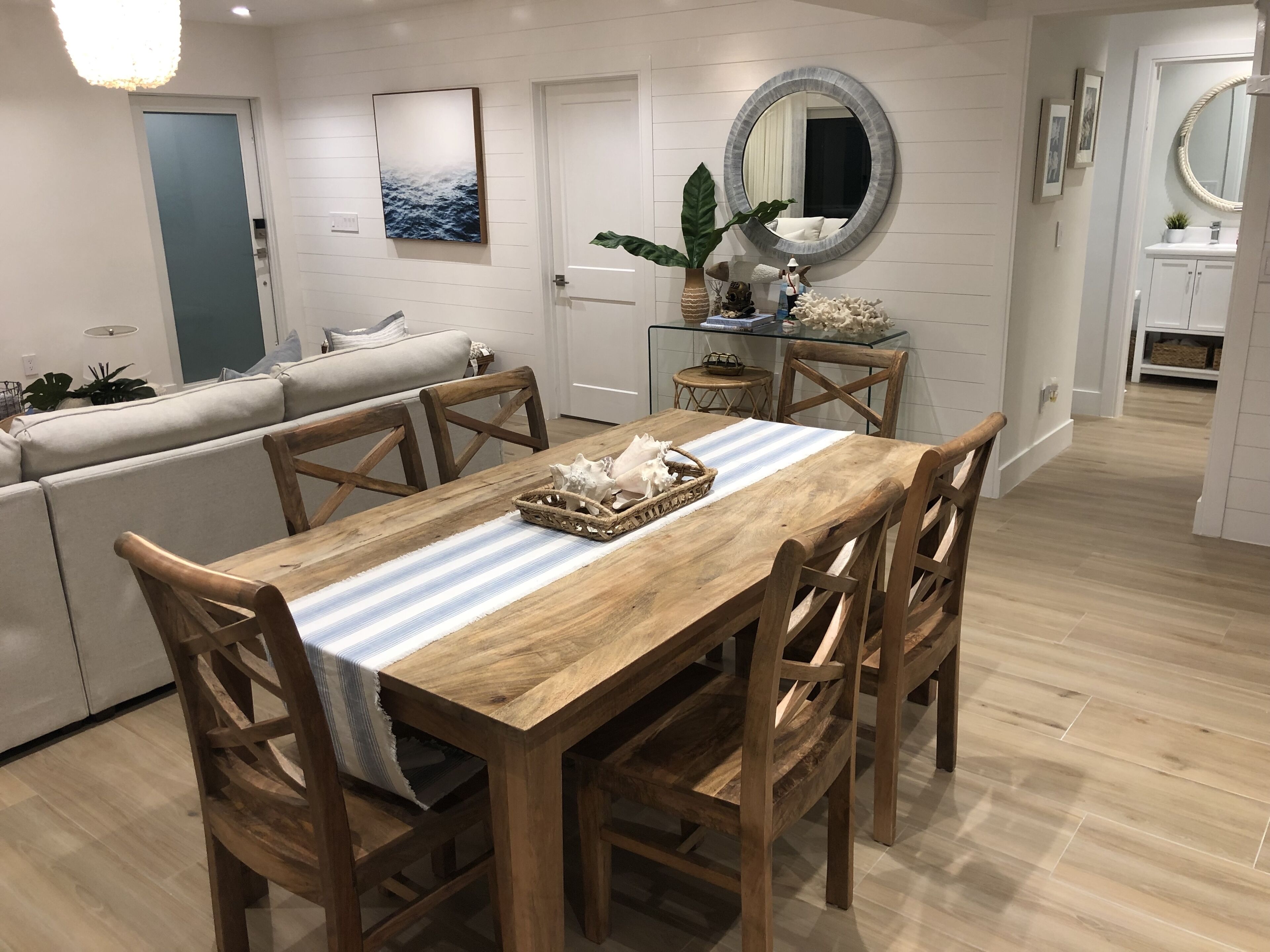 A dining area with a wooden table, chairs, and a striped tablecloth. There is a sofa with cushions in the background, a potted plant, and a mirror above the table.