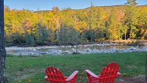 Zwei rote Adirondack-Stühle, die auf einen Fluss mit Wald im Hintergrund zeigen.