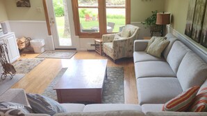 Ein Wohnzimmer mit einer Couch, einem Couchtisch und einem Sessel. Eine Tür führt zu einem Außenbereich mit einem Garten.