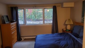 Ein Schlafzimmer mit einem Bett, einem Schminktisch, einem Nachttisch mit Lampe, einer Uhr und einem Fenster mit Vorhängen.