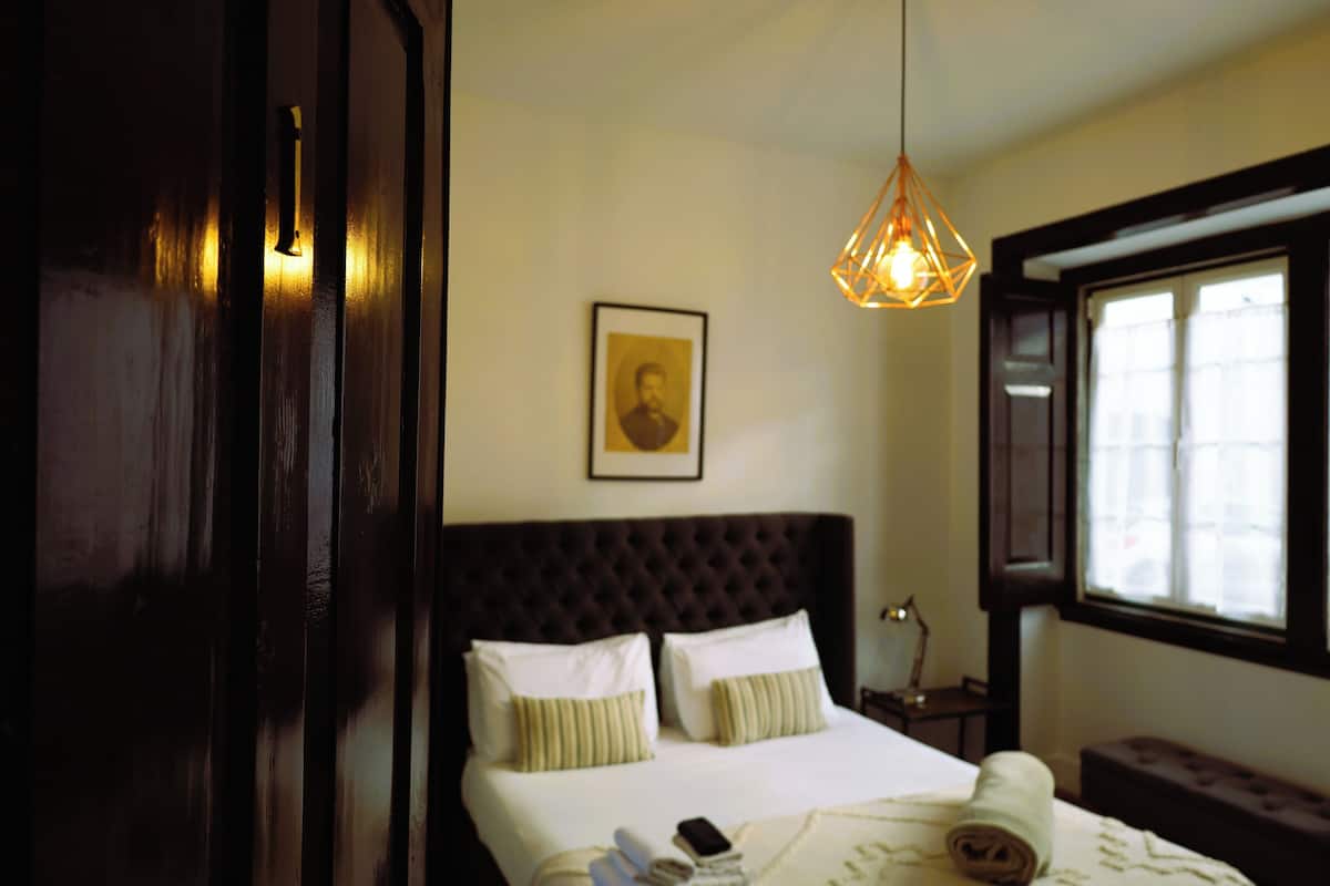A bedroom with a large bed, a hanging light fixture, a framed picture on the wall, and a window with blinds.