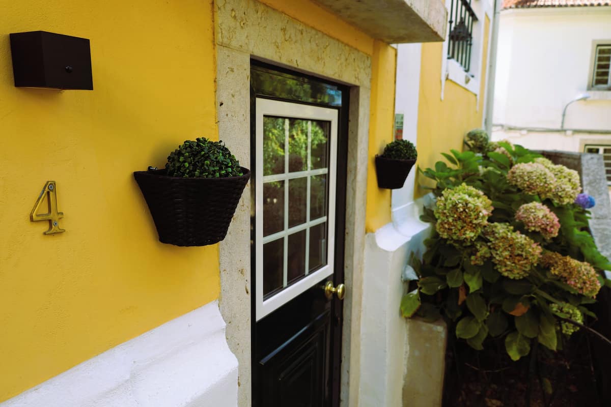A yellow building with a black door and window, a mailbox, and hanging flower baskets.