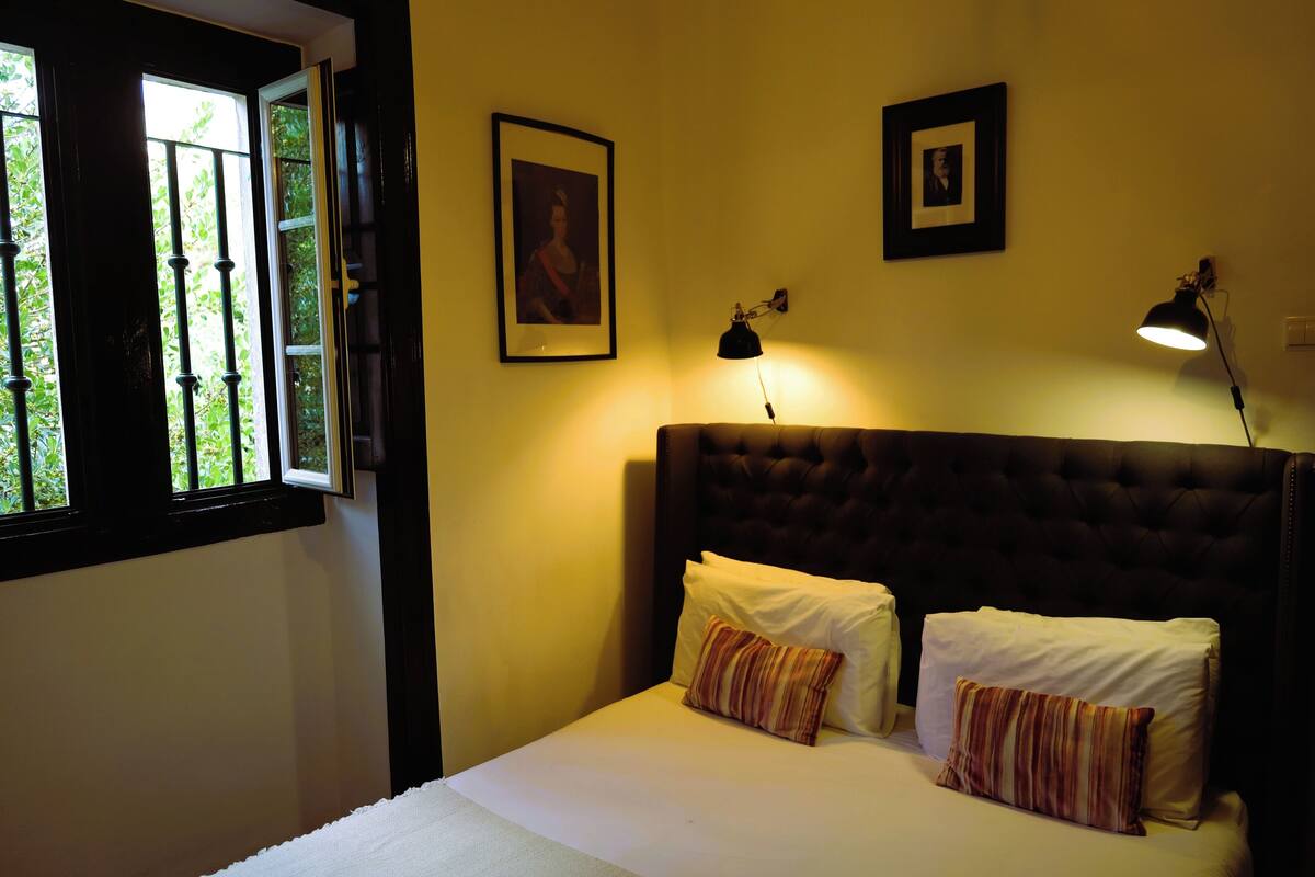 A bedroom with a bed, two pillows, a window with bars, and two wall-mounted lamps.