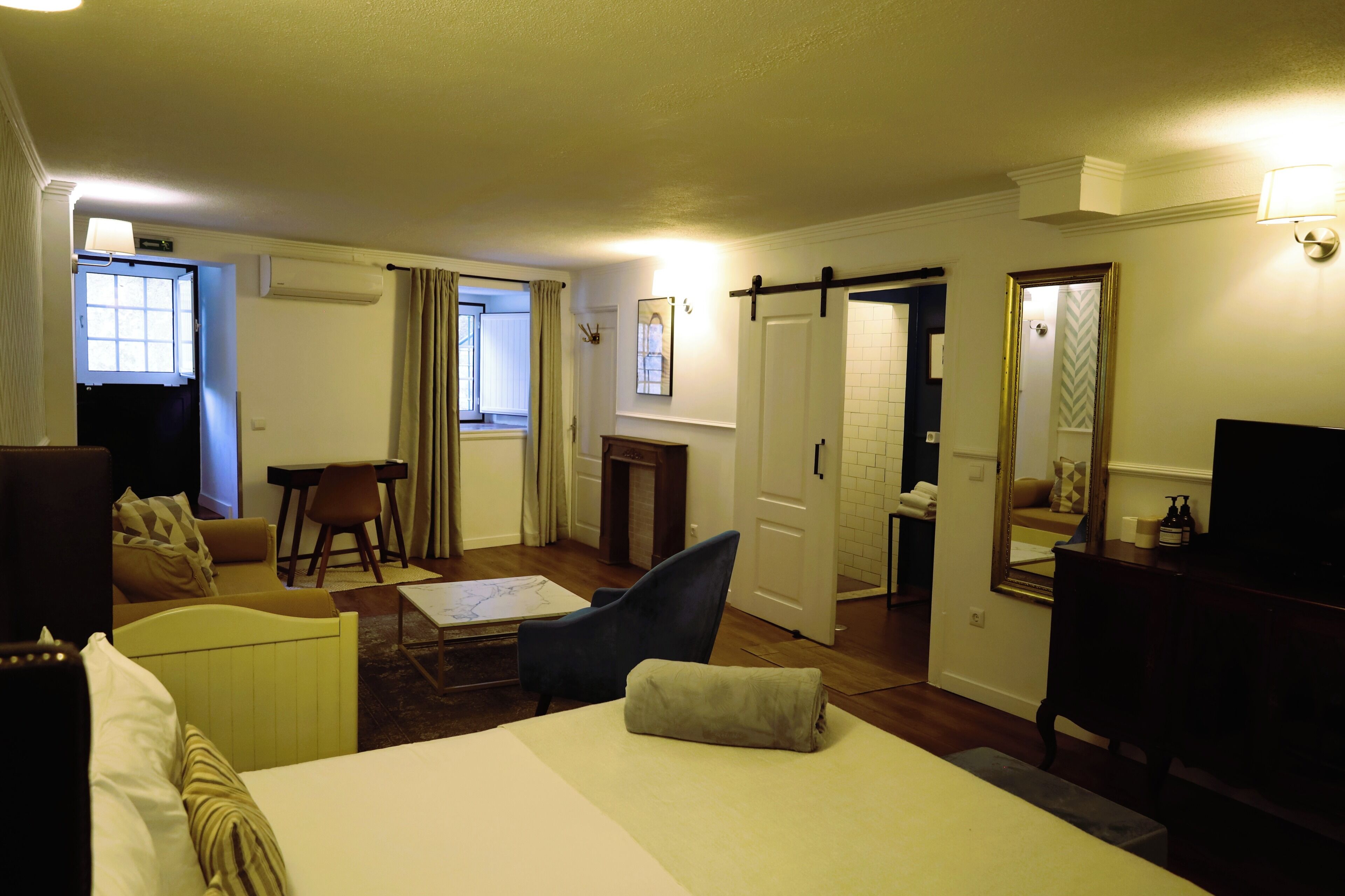 A hotel room with a bed, a desk, a chair, a sofa, a TV, and a bathroom visible through an open door.