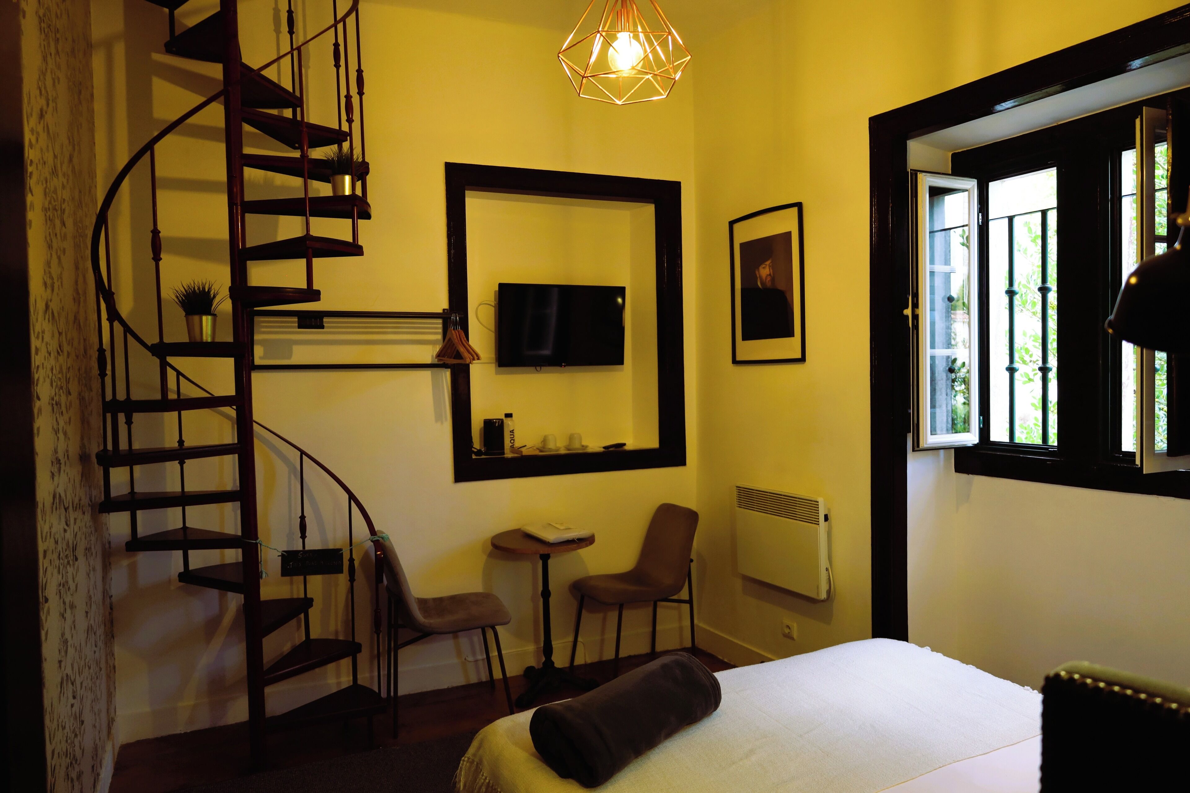 A bedroom with a bed, a chair, a small table, a TV, a window, and a spiral staircase.