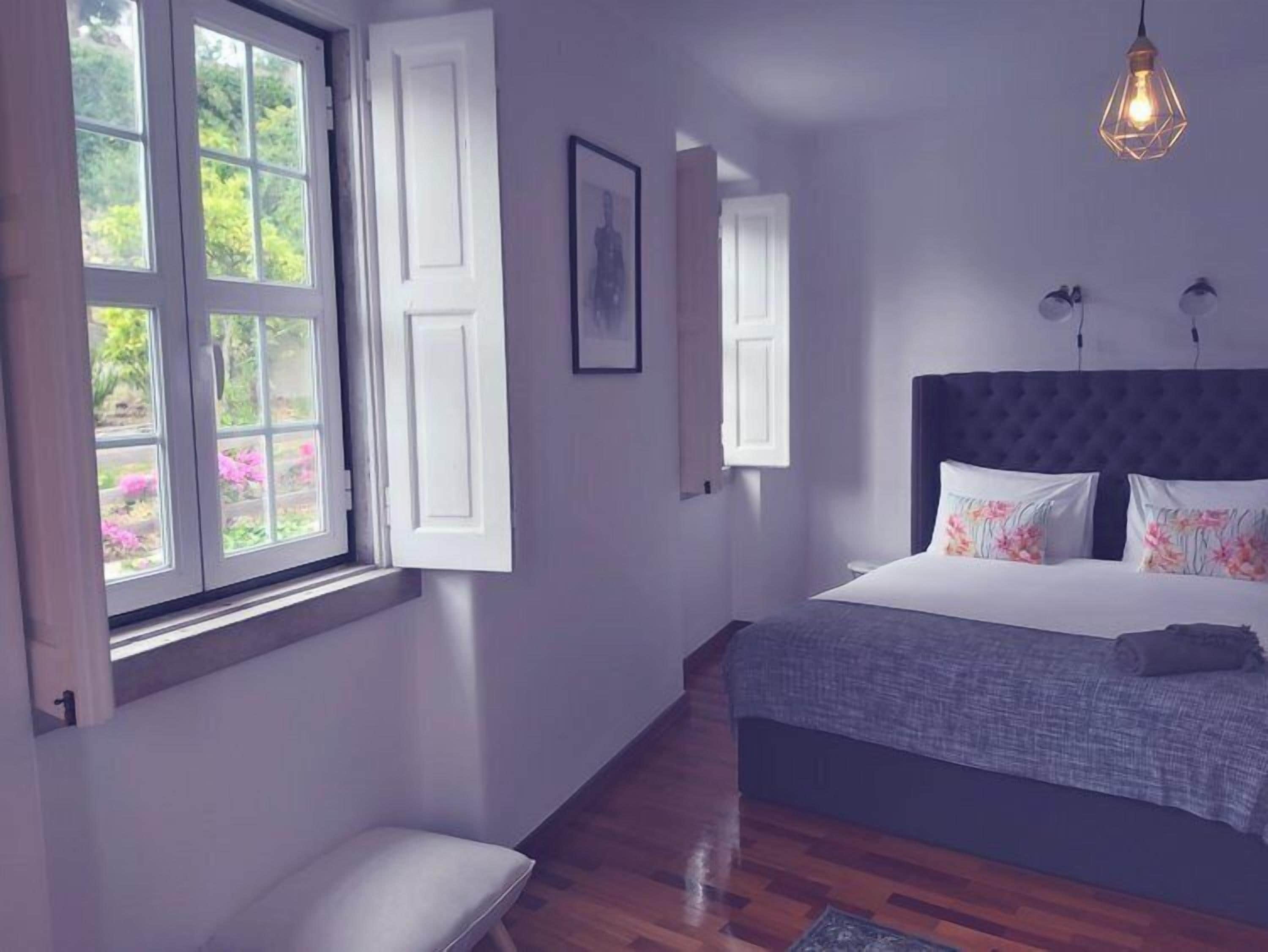 A bedroom with a large bed, a window with open shutters, and a hanging light fixture.