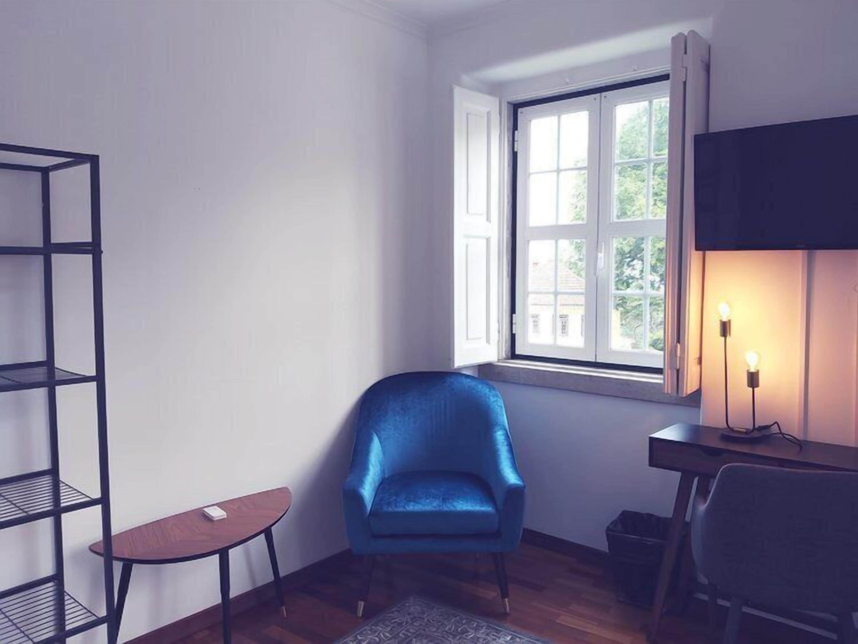 Una habitación en esquina con una silla azul, una mesita, una estantería, una ventana con cortinas y un escritorio con lámpara.