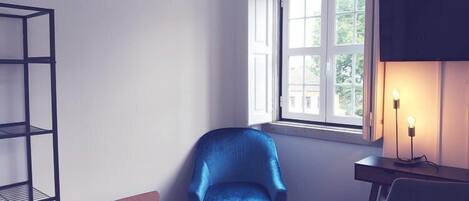 Una habitación en esquina con una silla azul, una mesita, una estantería, una ventana con cortinas y un escritorio con lámpara.