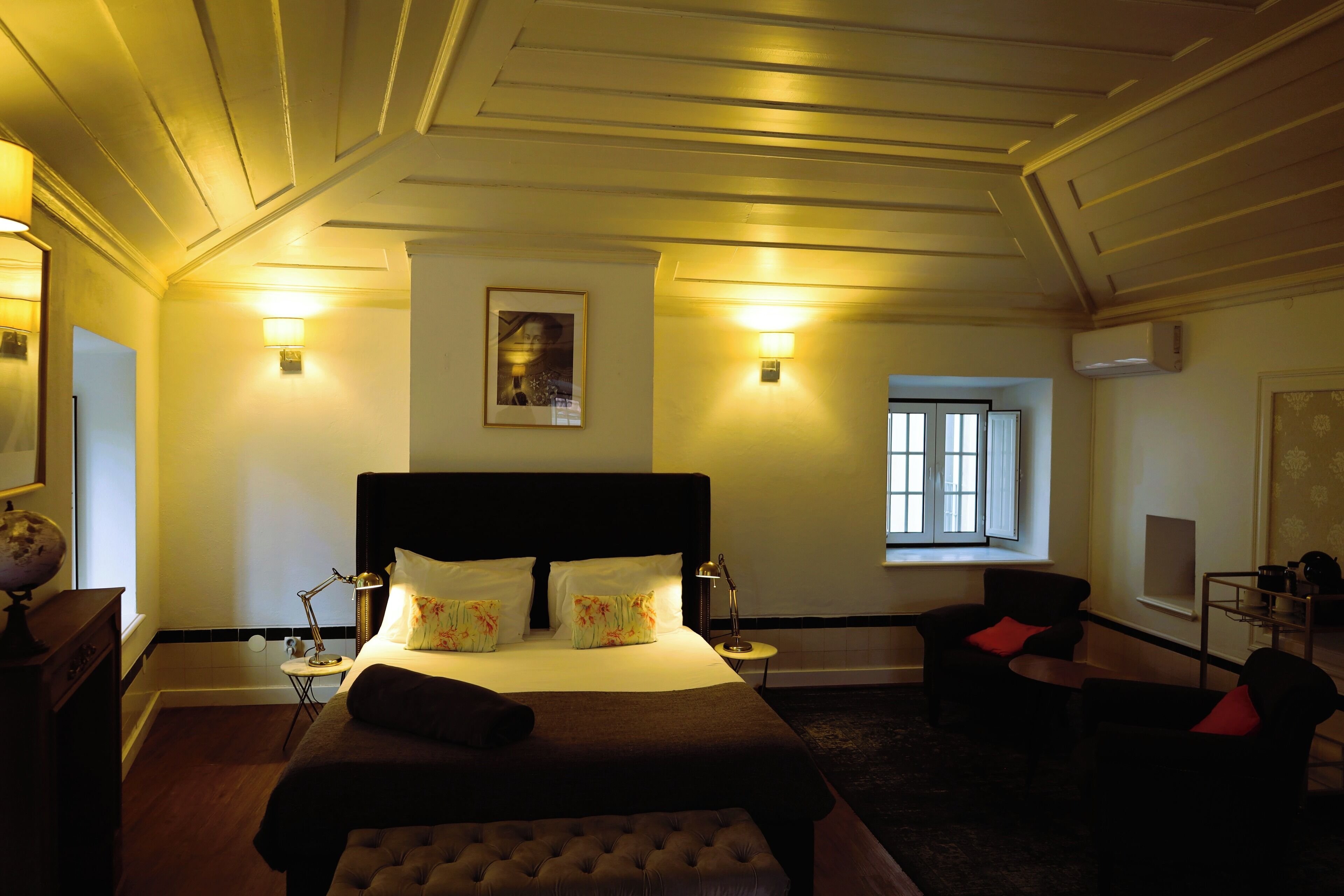 A hotel room with a large bed, bedside lamps, a small table, a chair, and a window.