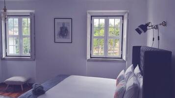 Une chambre à coucher avec un lit, deux fenêtres, un tableau encadré, un petit tabouret et une lampe.