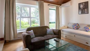 A living room with a brown sofa, a glass coffee table, and a white sofa with colorful pillows. There are two windows with curtains, and an air conditioning unit on the wall.
