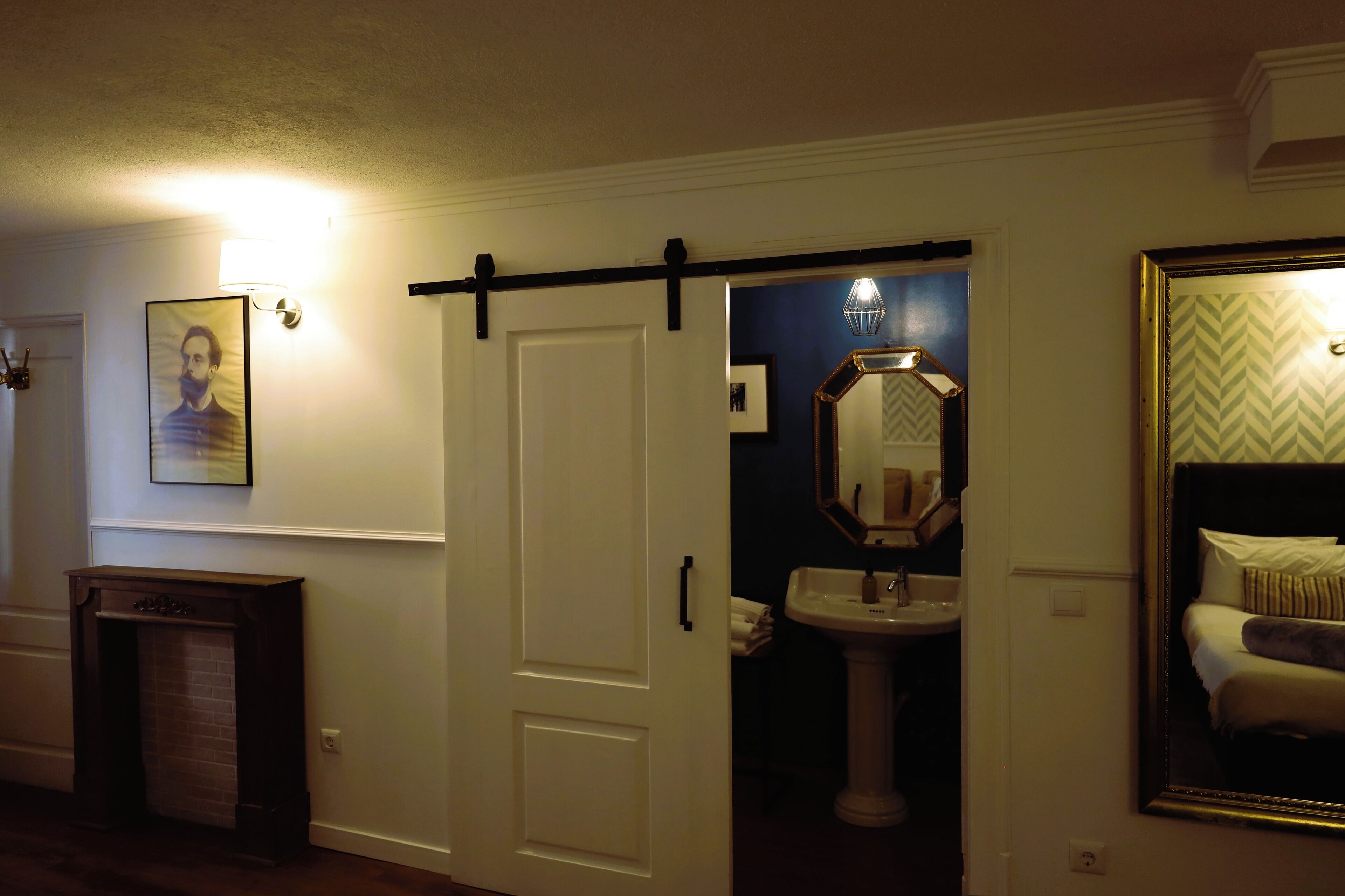 A hotel room with a sliding door, a white wall, a framed picture, a mirror with a geometric pattern, and a bed with white bedding.