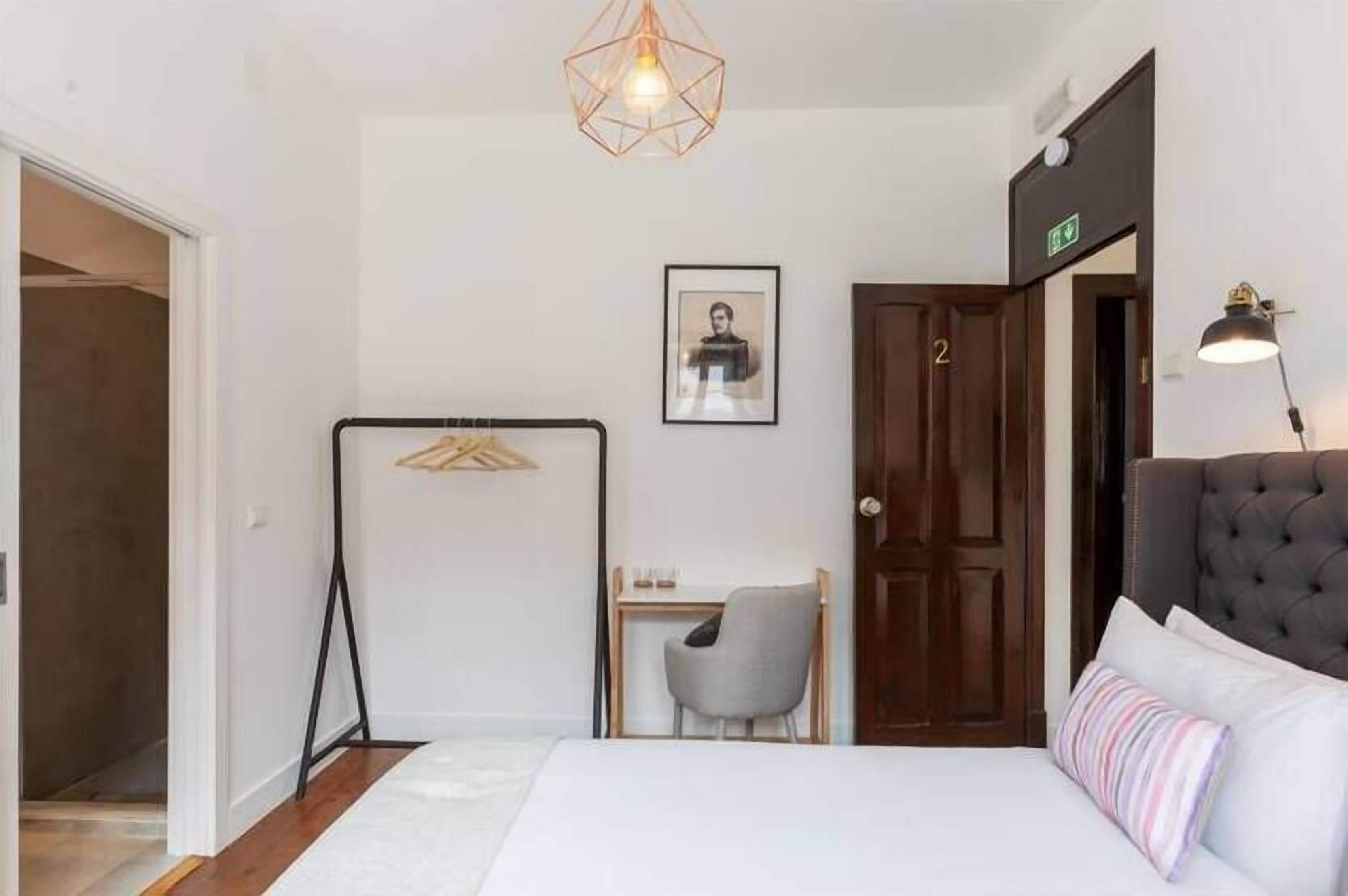 A hotel room with a bed, a desk, a chair, a hanging light fixture, and a door with an exit sign.