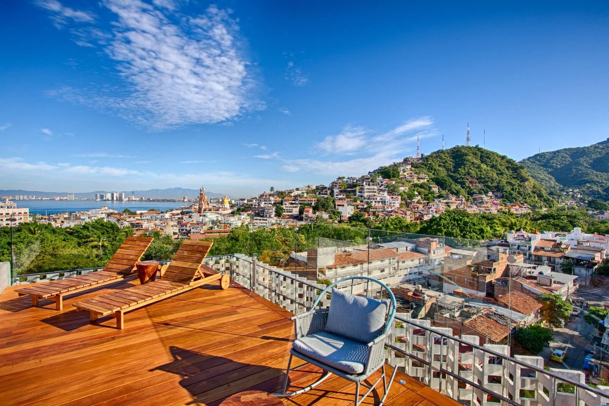 Terraza en la azotea con piso de madera, sillones de descanso y vistas a una ciudad costera.