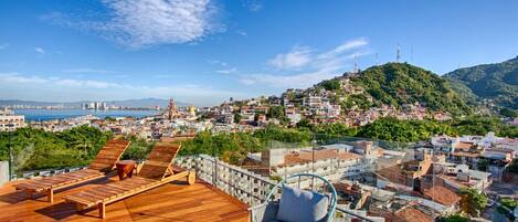 Terraza en la azotea con piso de madera, sillones de descanso y vistas a una ciudad costera.