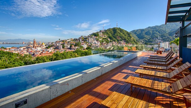A rooftop infinity pool with wooden deck and lounge chairs, overlooking a cityscape with buildings and hills. - Chic, Spacious Condo | Great Walkability, Pool, Gym, Rooftop Views (Puerto Vallarta)