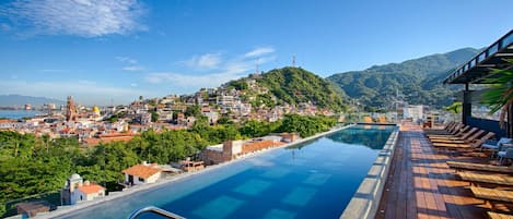 Une piscine à débordement située sur le toit, avec une terrasse en bois et des chaises longues, offrant une vue sur une ville côtière et les collines environnantes.