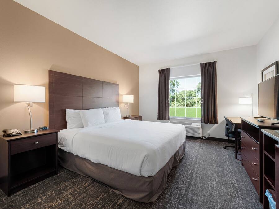 A hotel room with a large bed, two bedside tables with lamps, a desk with a chair, a TV, and a window with curtains.