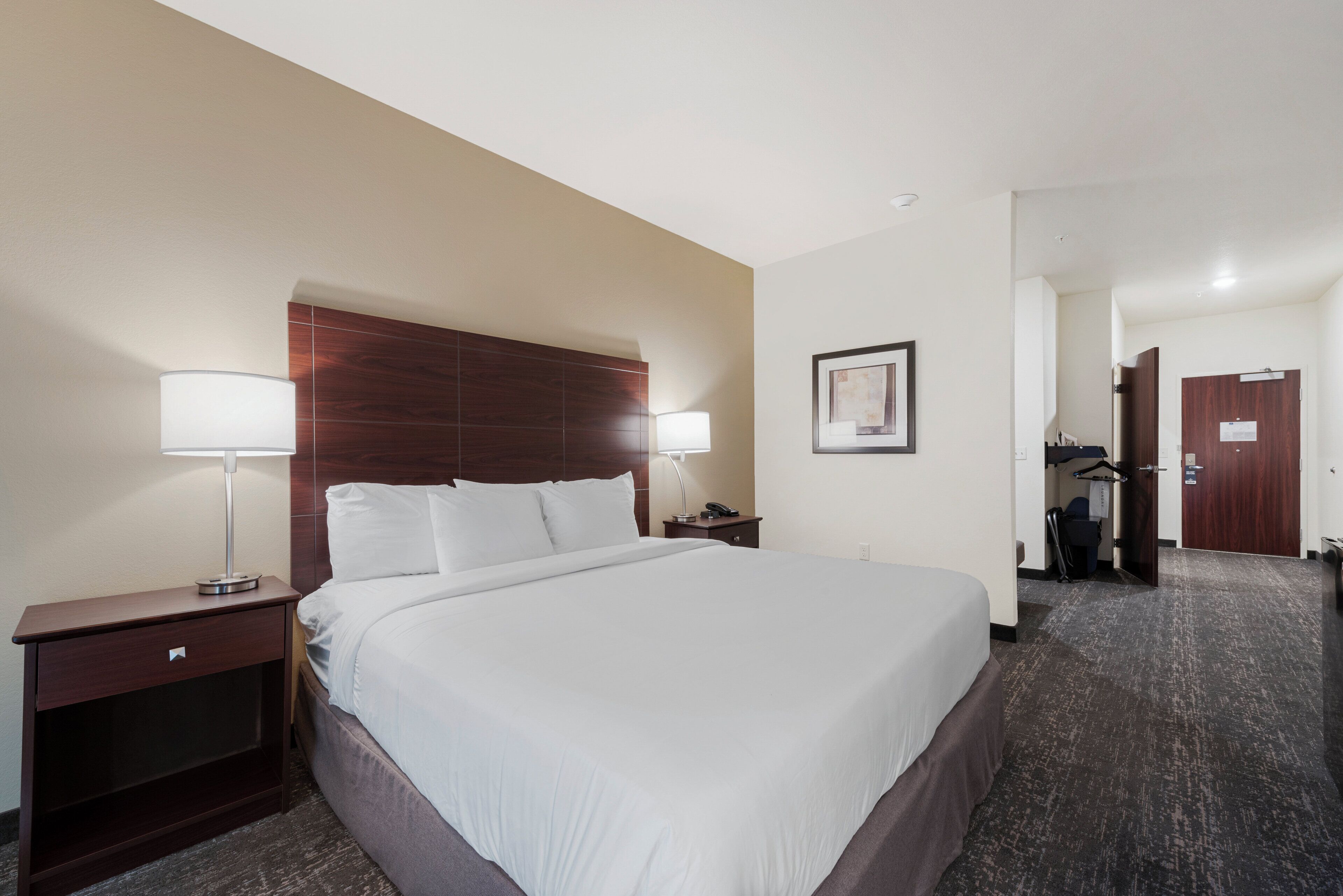A hotel room with a large bed, two bedside tables with lamps, a wooden headboard, a framed picture on the wall, and a door leading to another room.