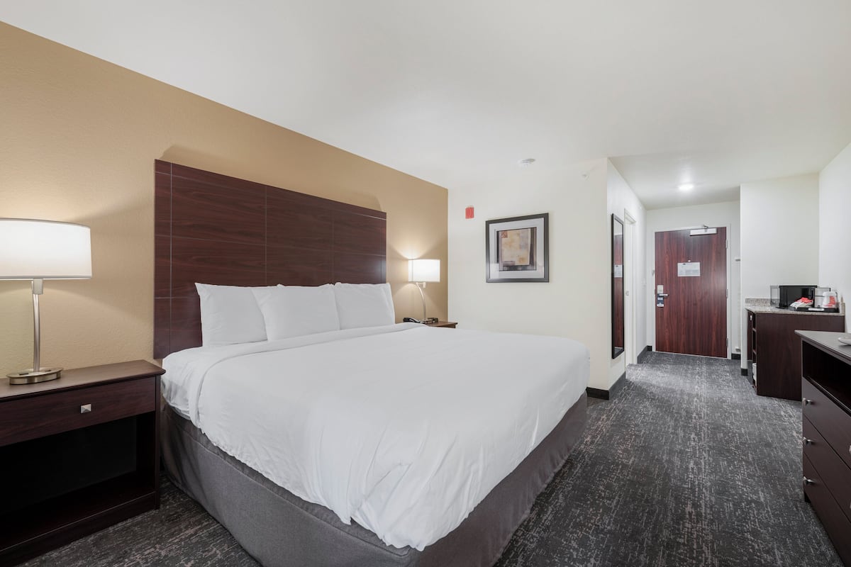 Une chambre d’hôtel avec un grand lit, une table de chevet, un bureau, une chaise, une lampe, un tableau au mur et une porte donnant sur une autre pièce.