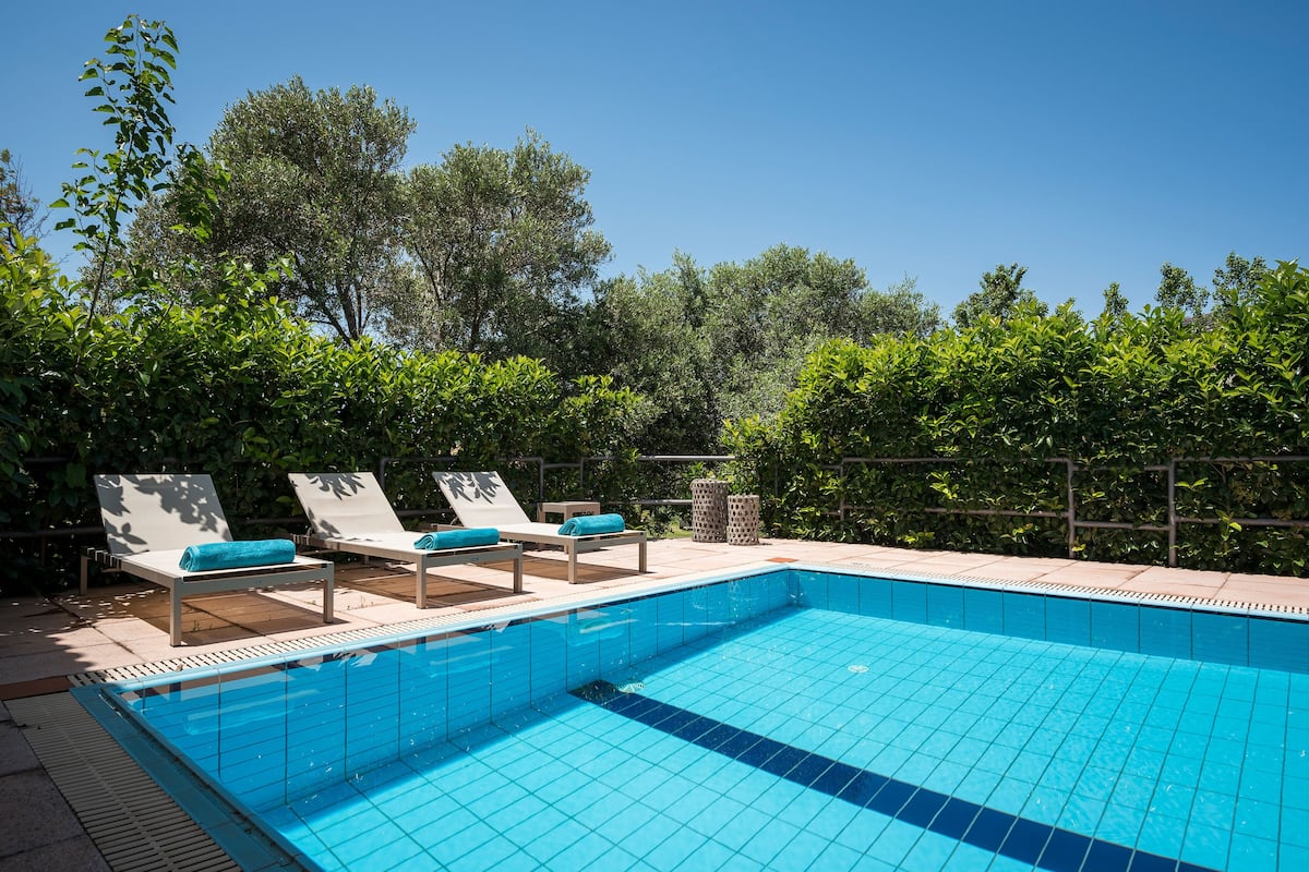 A swimming pool with tiled blue floor, surrounded by lounge chairs with blue cushions, and a hedge in the background.
