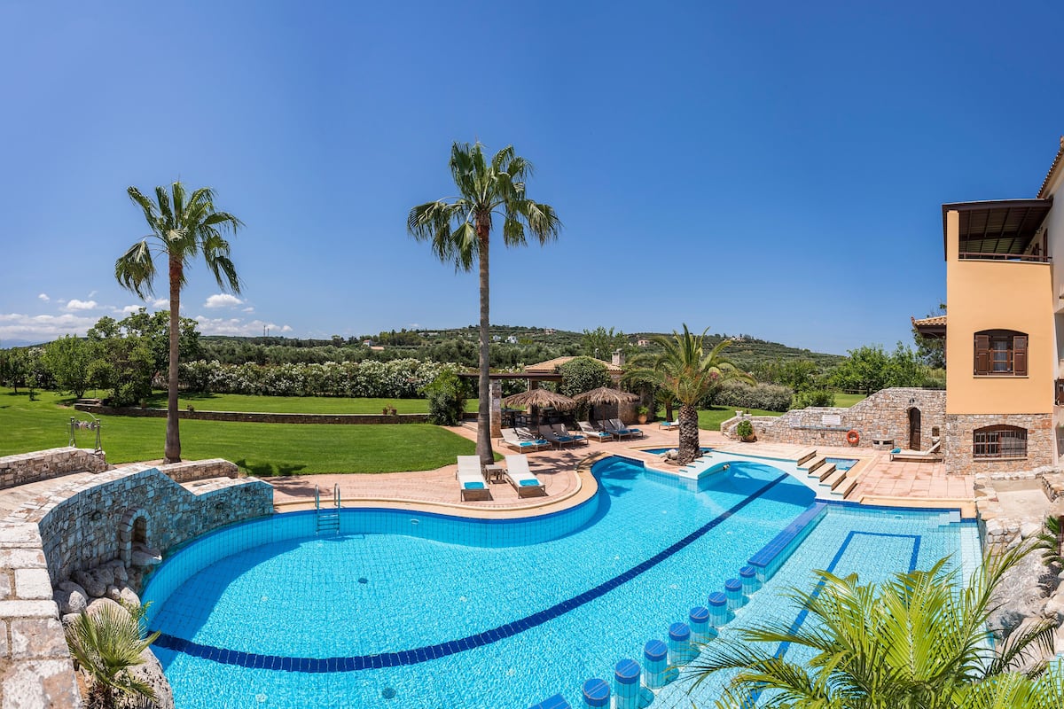 A large outdoor swimming pool with a curved design, surrounded by lounge chairs and palm trees, set against a backdrop of a hilly landscape and a clear blue sky.