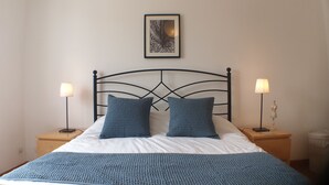 Un lit bien fait, recouvert d’une couverture bleue tricotée et agrémenté d’oreillers, avec deux tables de chevet de chaque côté, chacune surmontée d’une lampe. Le tout est placé contre un mur sobre, au-dessus duquel est accrochée une image encadrée.