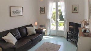 A living room with a dark leather sofa, a TV, and a small table with a lamp. There are two windows with curtains, and a door leading outside. - Lavender Cottage is a one bedroom gîte with private terrace and garden (Étaules, Nouvelle-Aquitaine,)