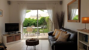 Una sala de estar con un sofá de cuero negro, televisor, una mesa de comedor con sillas y vista a un jardín a través de un ventanal.