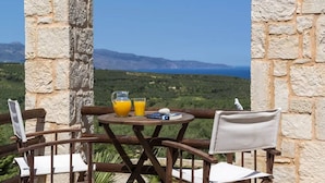 Ein Tisch mit einem Glas Orangensaft, einem Buch und einer Fernbedienung. Der Tisch steht auf einem Balkon mit Blick auf das Meer und die Berge.