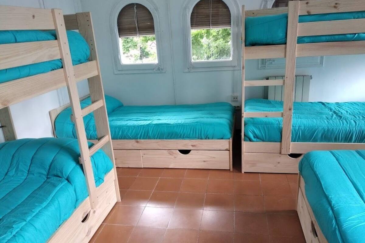 A room with four bunk beds, each with a wooden frame and teal bedding. There are two windows with arched tops, and the room has a tiled floor.
