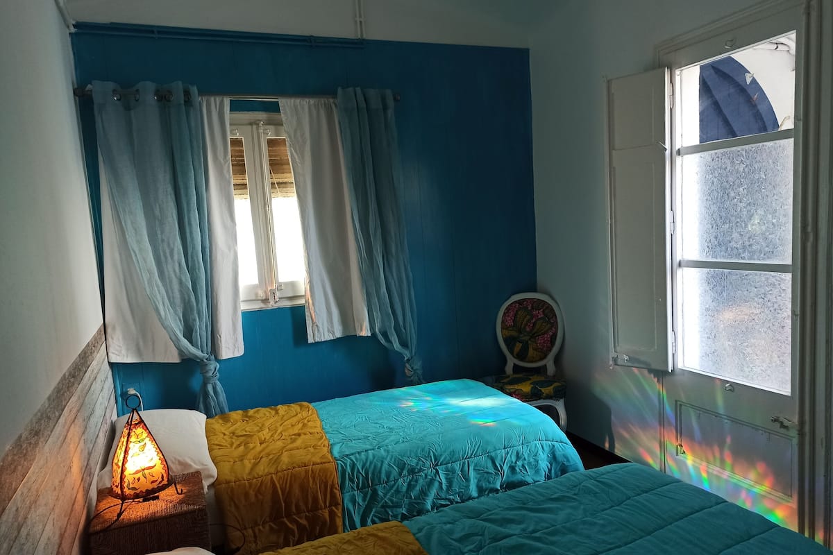 A bedroom with two beds, a window with curtains, a nightstand with a lamp, and a chair with a floral headrest.