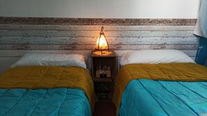 Una habitación con dos camas, cada una con una colcha amarilla y azul. Entre las camas, un mesita de noche con una lámpara.
