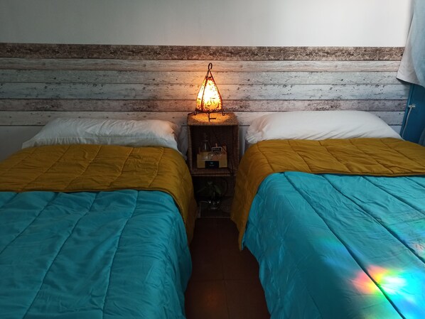 Una habitación con dos camas, cada una con una colcha amarilla y azul. Entre las camas, un mesita de noche con una lámpara.