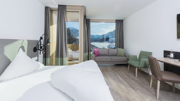 Habitación de hotel con cama, escritorio, silla y sofá. Se ve una vista de montañas y un lago por la ventana.