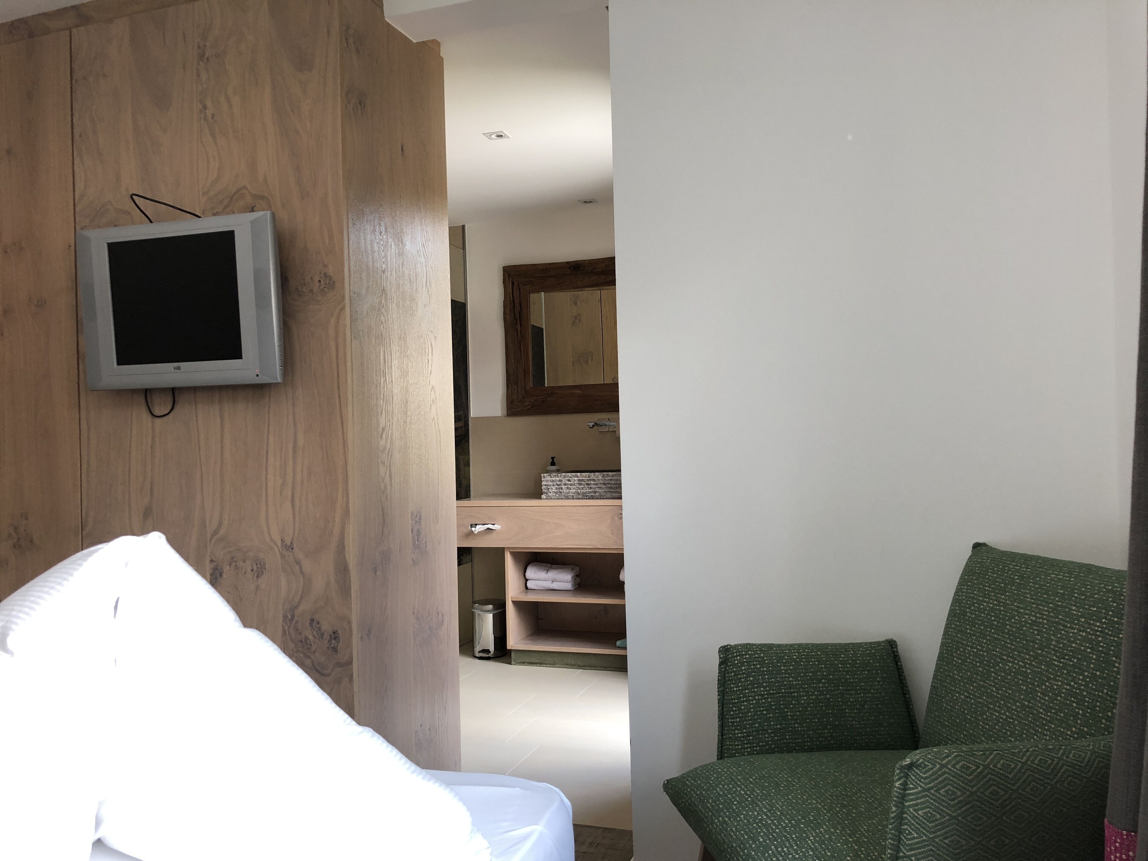 Un dormitorio con cabecera de madera, una mesita de noche, una silla verde con estampado, un televisor montado en la pared y un baño visible a través de una puerta abierta.