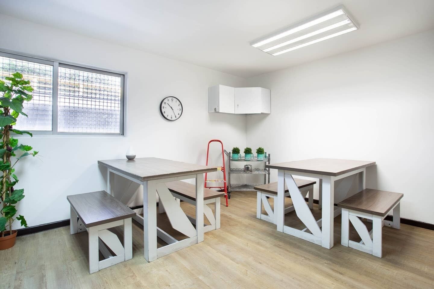 Una habitación moderna y luminosa con piso de madera, una mesa de comedor con bancos, una silla roja, un reloj, una ventana con persianas y una planta en maceta.