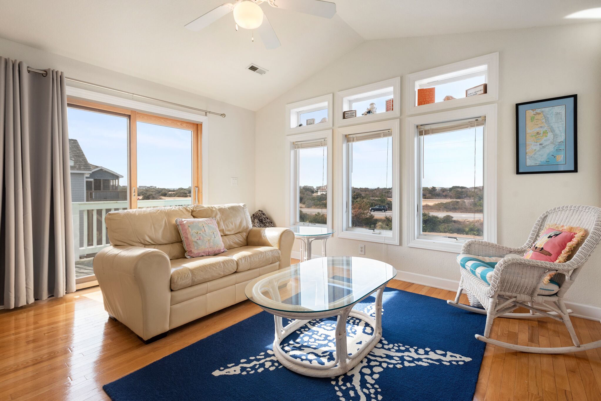 A spacious living room with a glass-top coffee table, a beige sofa, a rocking chair, and large windows offering a view of the landscape.