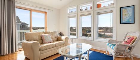 A spacious living room with a glass-top coffee table, a beige sofa, a rocking chair, and large windows offering a view of the landscape.