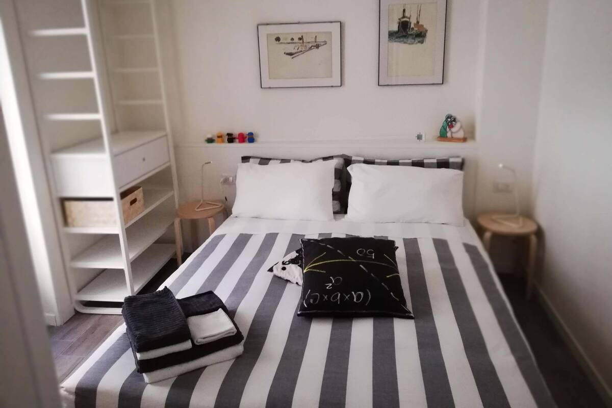 Quarto bem arrumado com colcha listrada, travesseiro preto e branco e uma pequena mesa de cabeceira com abajur. Há duas fotos emolduradas na parede, um guarda-roupa branco e um conjunto de garrafas coloridas em uma prateleira.