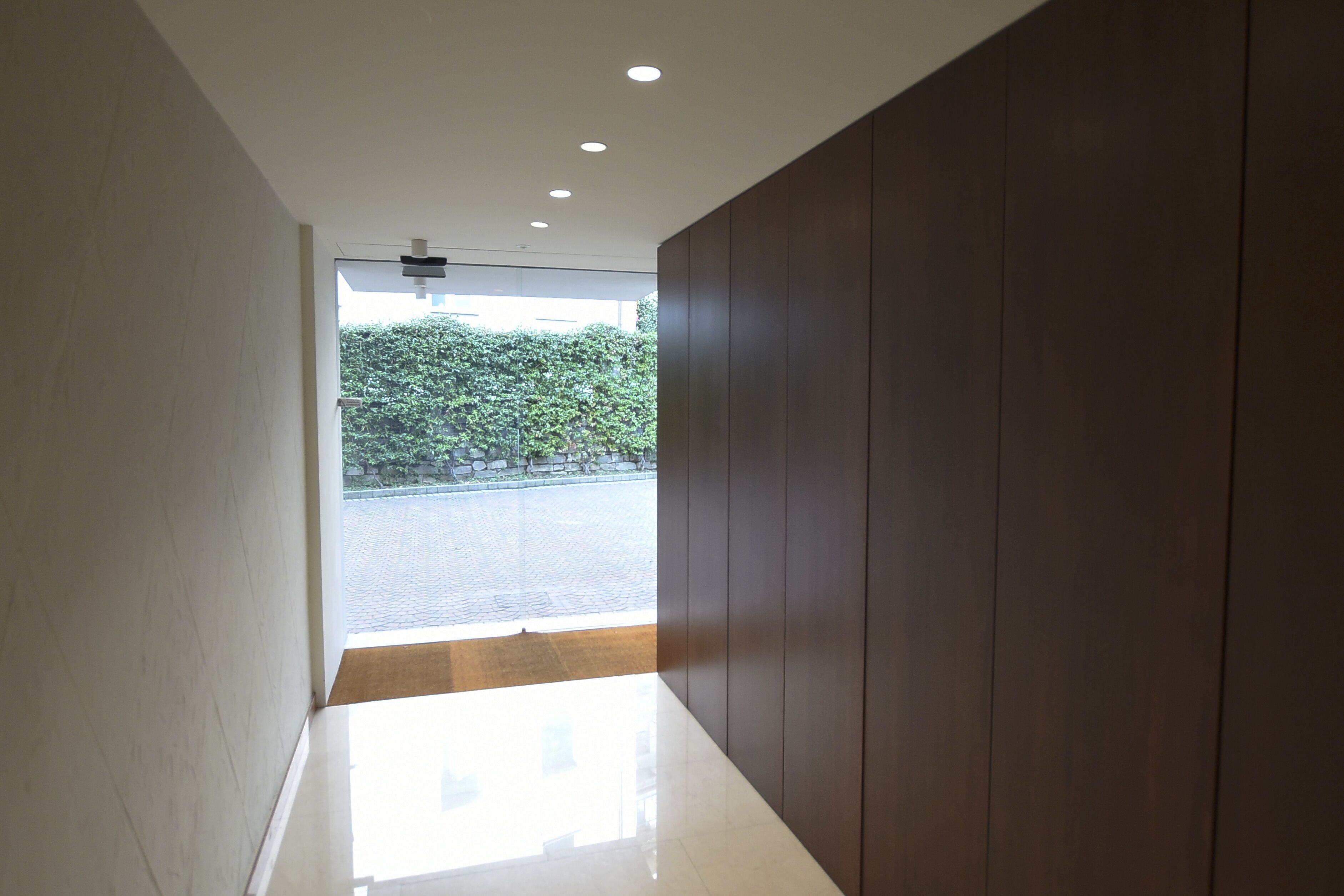 A modern hallway with a polished floor, recessed ceiling lights, and a view of a paved area and greenery outside.