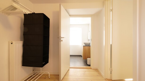 En smal hall med ett element, ett svart föremål på ett ställ och en öppen dörr som leder till ett badrum.