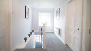 A hallway with white walls, a grey carpet, and a white door at the end. There are small potted plants on the left side.